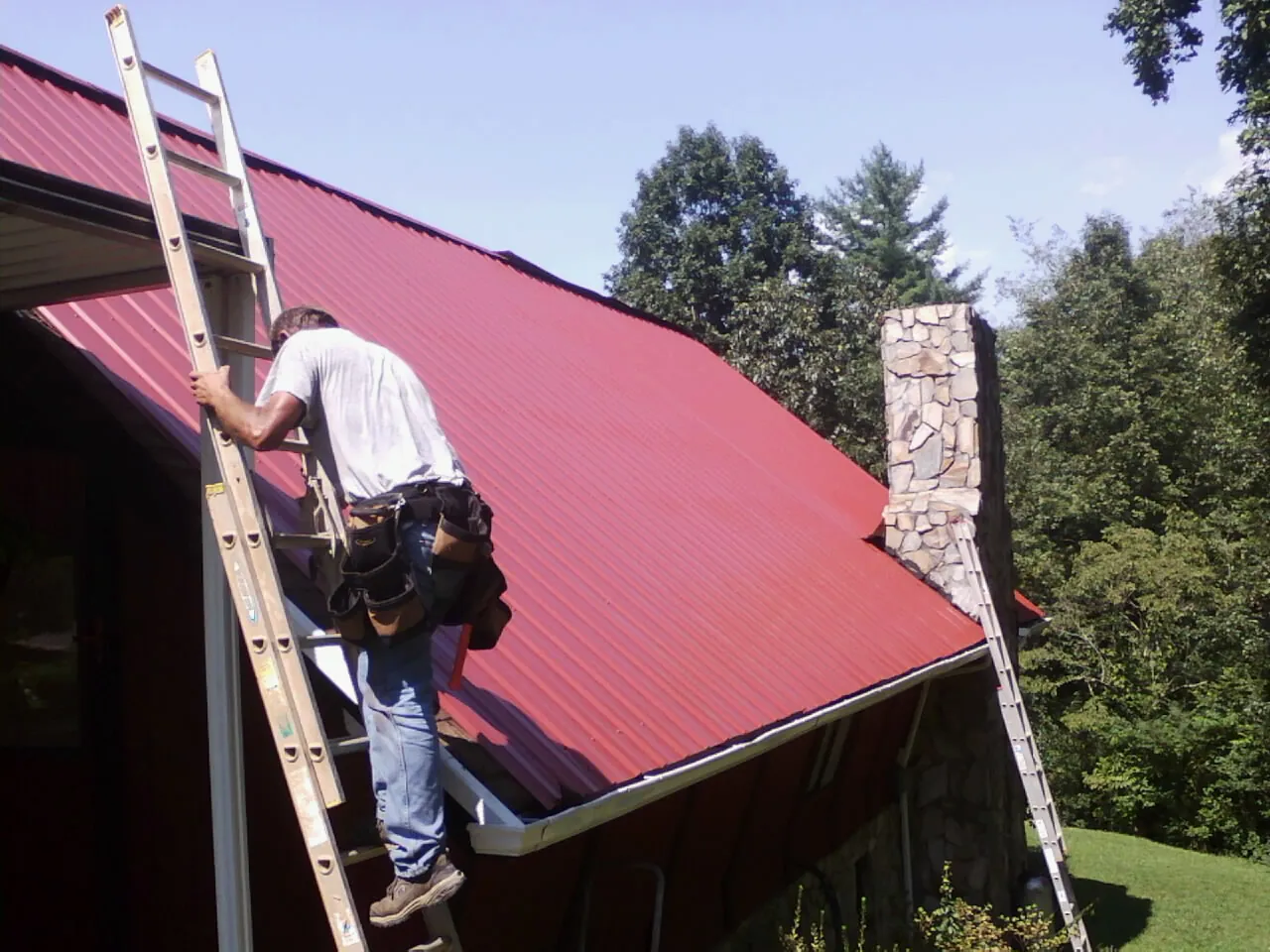 Roofer on a ladder beside a red metal roof and stone chimney — Soffit & Fascia Repair by Roofing Brothers in Jessieville